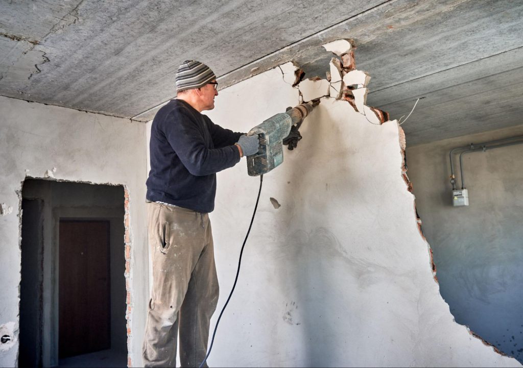 Aannemer breekt met een boorhamer een bakstenen muur uit tijdens een renovatie, met een betonnen plafond zichtbaar op de achtergrond.