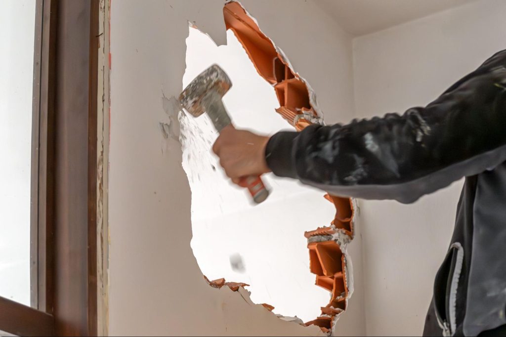 Arbeider breekt een binnenmuur open met een voorhamer, waarbij de oranje holle blokken zichtbaar worden.